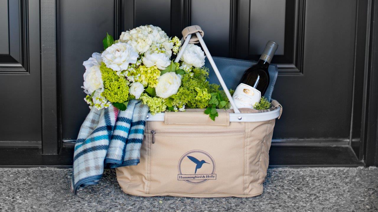 gift basket includes flowers, a throw blanket, bottle of red wine, in a picnic basket