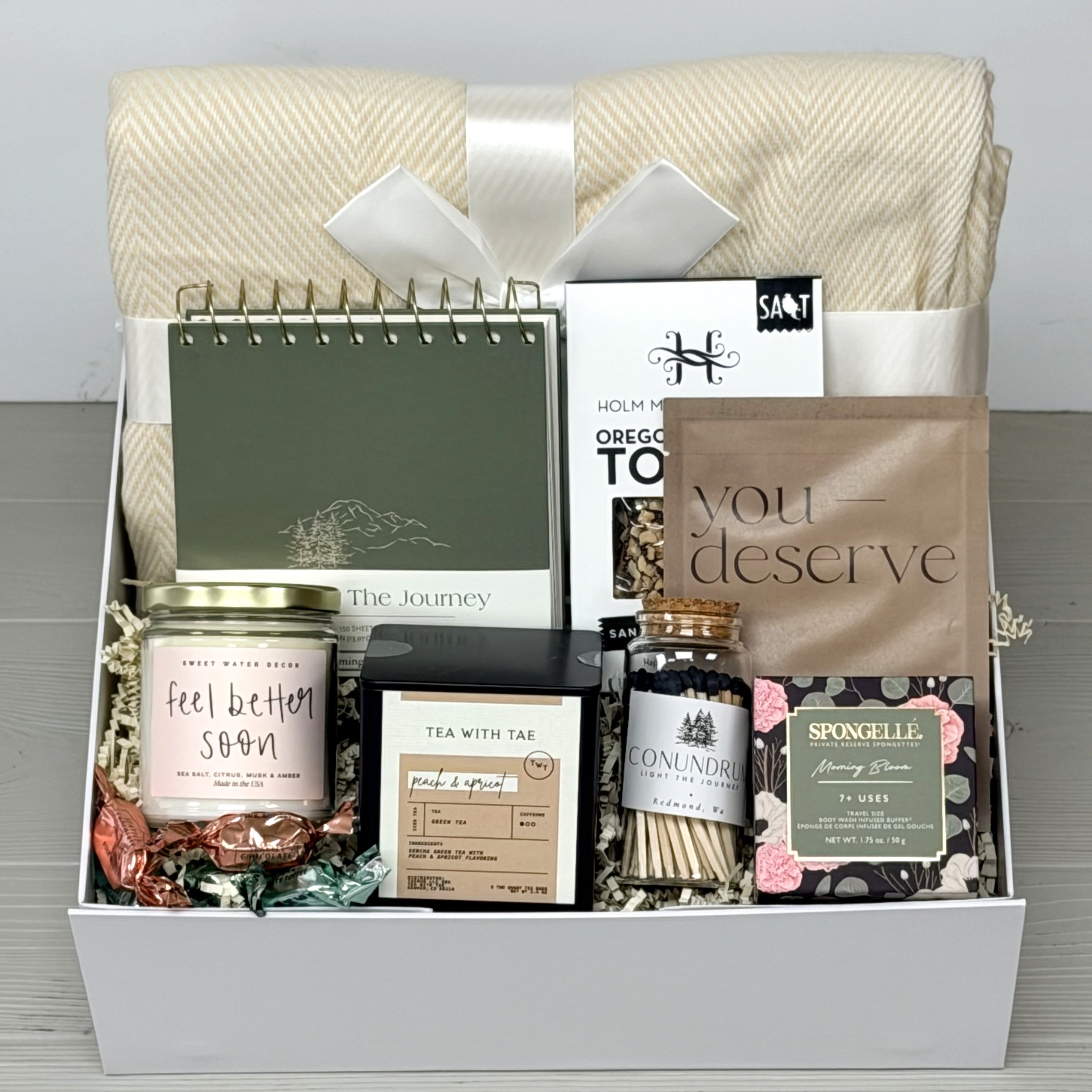 Gift basket with various items including a candle, tea, and a book on a gray surface.