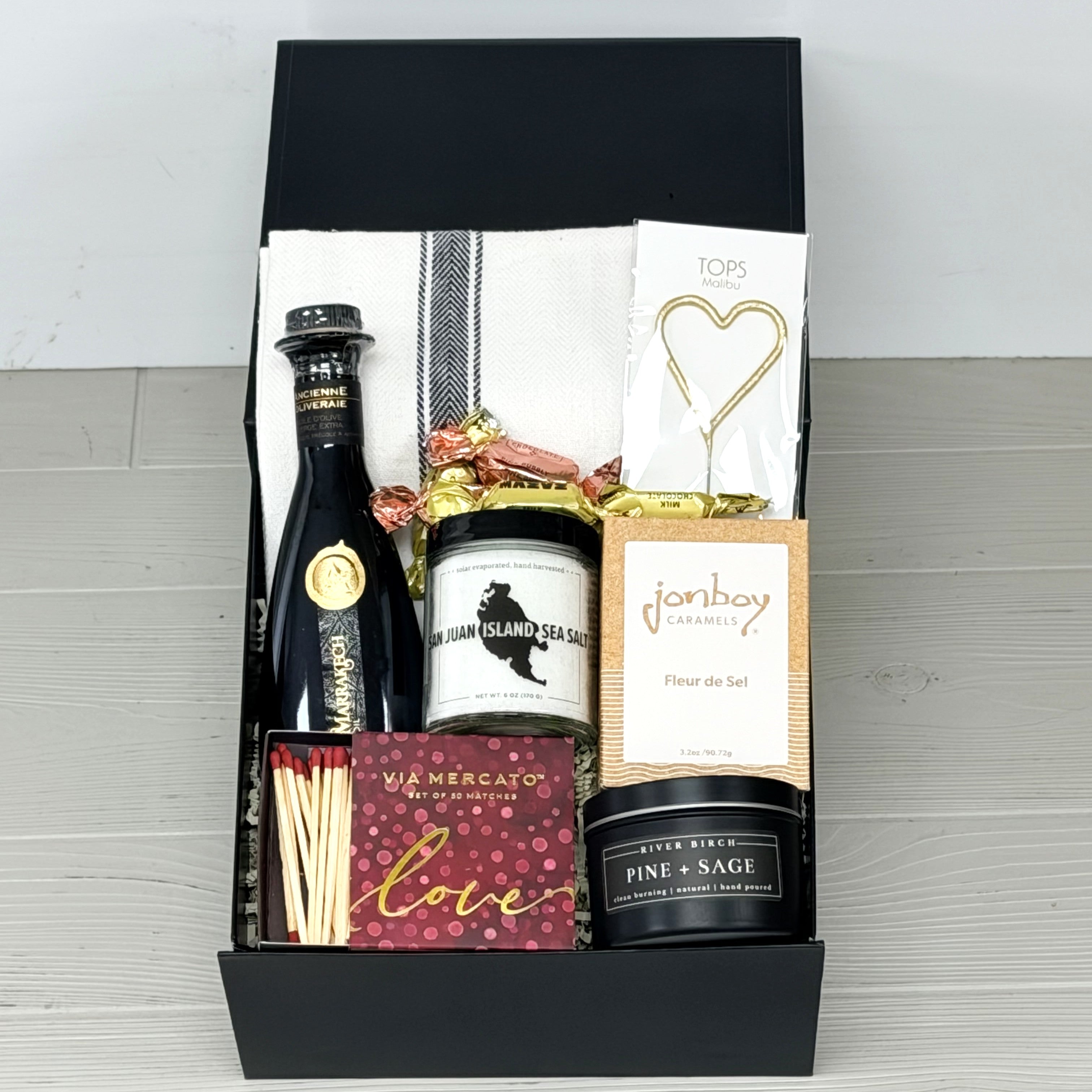 gift basket that contains olive oil, sea salt, caramel, candle, matches, candy, kitchen towel