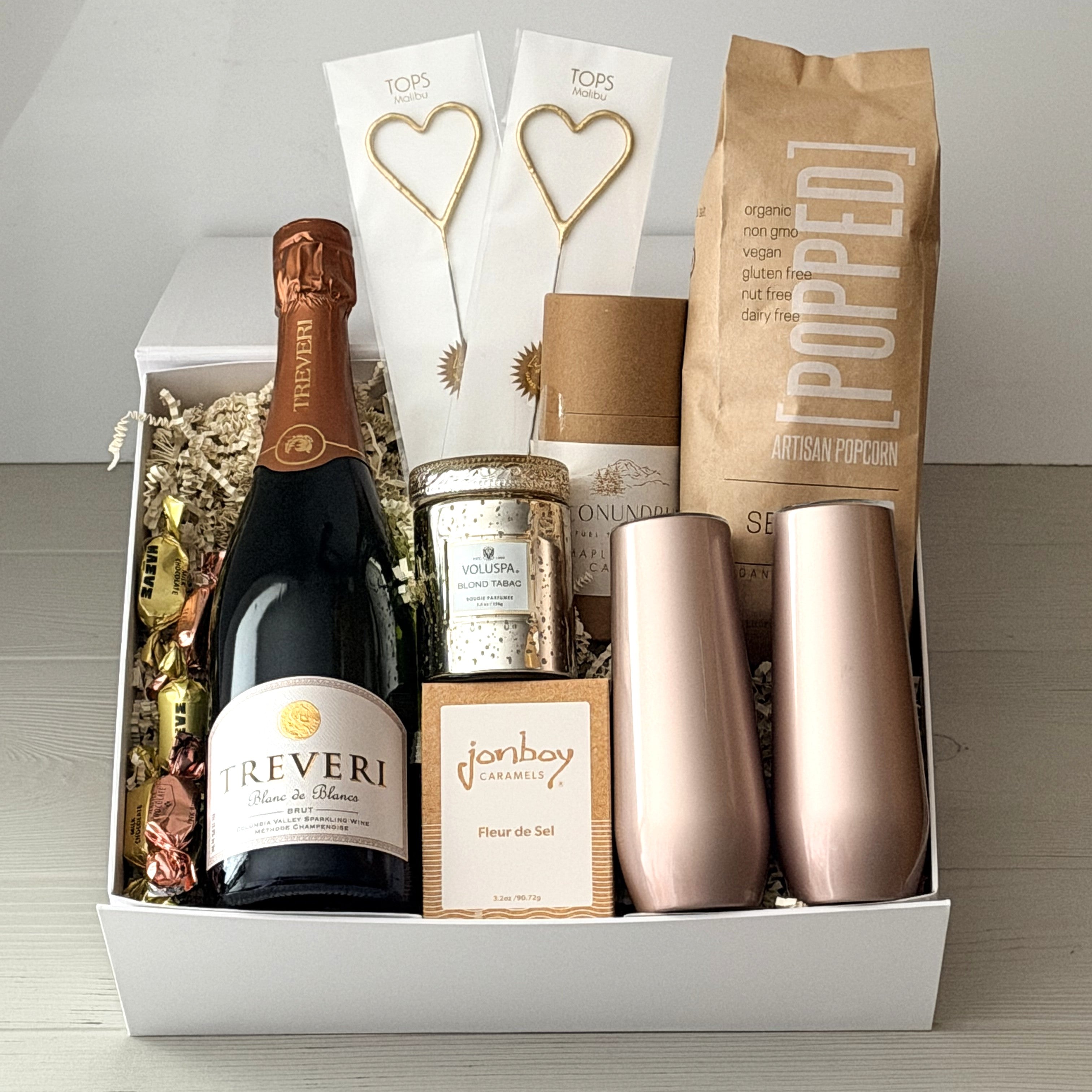 Gift basket with Treveri sparkling wine, rose gold tumblers, and other items on a wooden surface.
