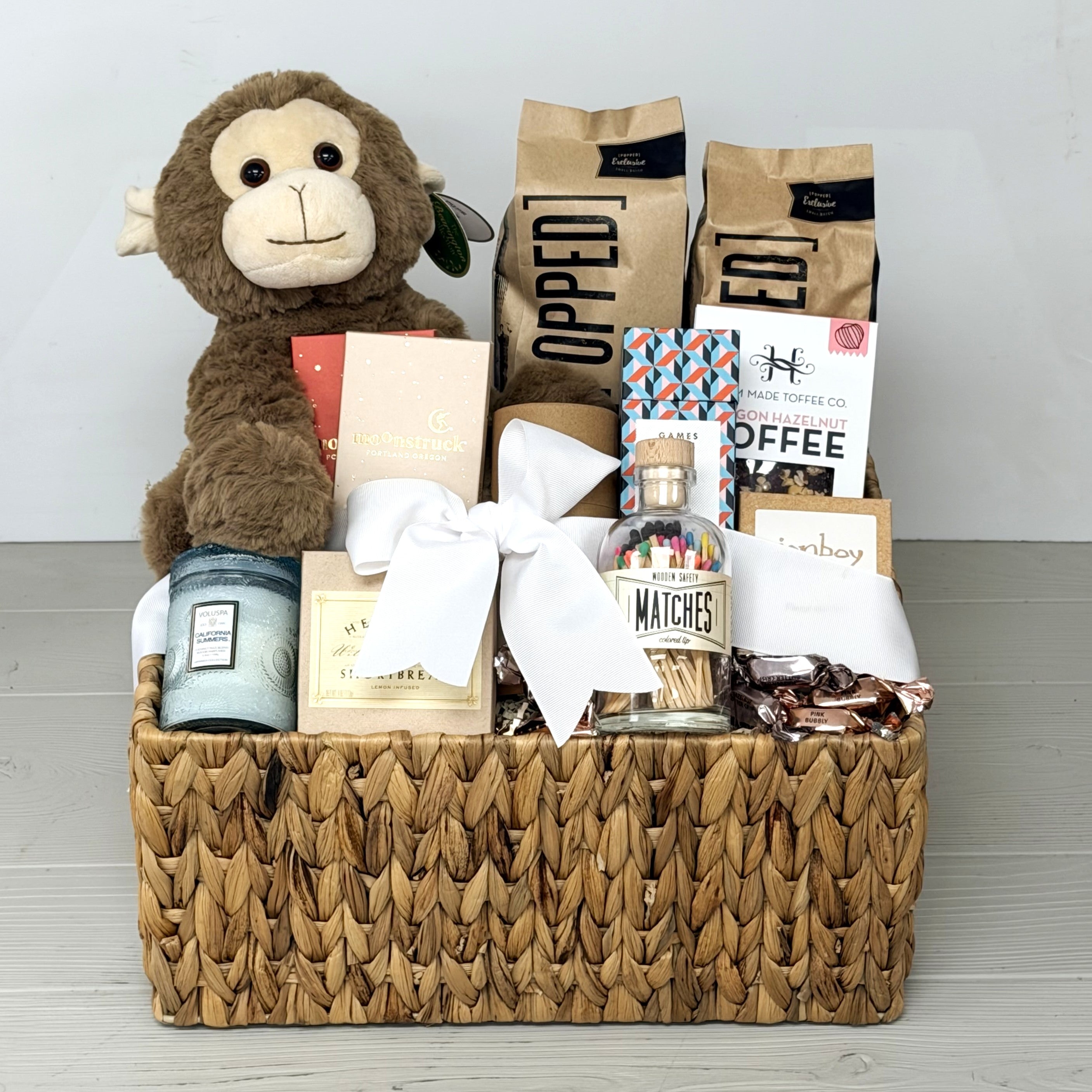 gift basket with plush monkey, popcorn, toffee, chocolate, candle, cookes, in a basket on light surface