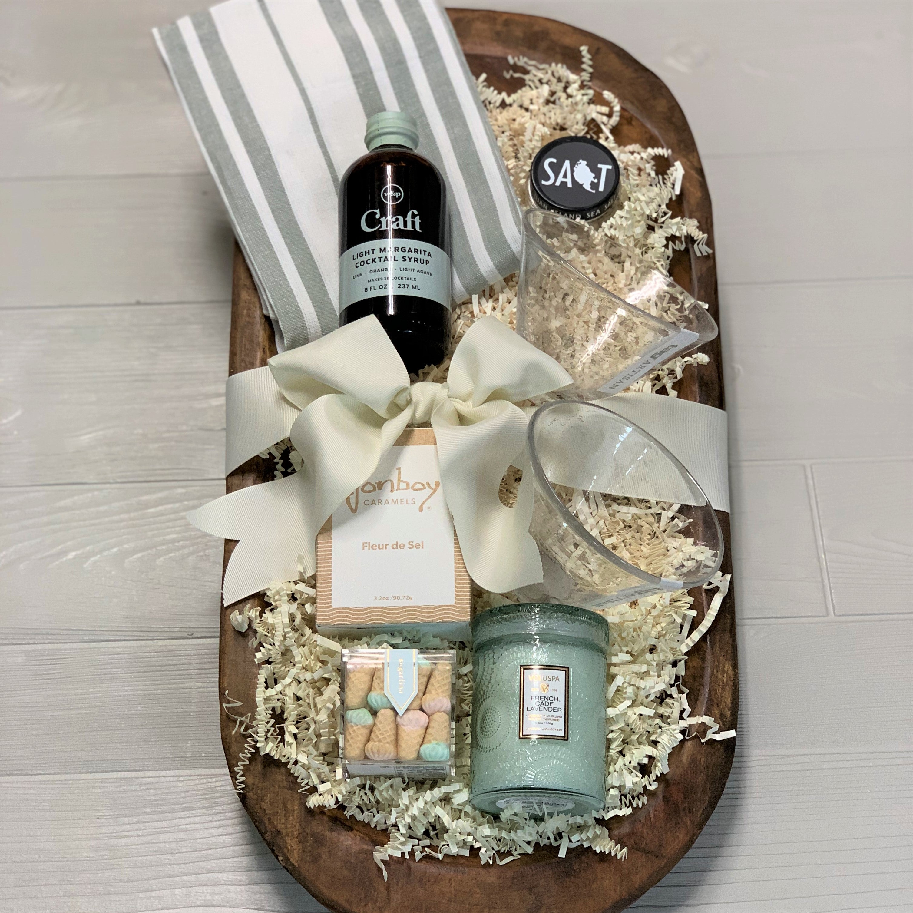 a margarita kit gift basket that includes a kitchen towel, margarita syrup, 2 glasses, caramel, Sugarfina gummy bears, sea salt, Voluspa candle, packaged in a wooden bowl.