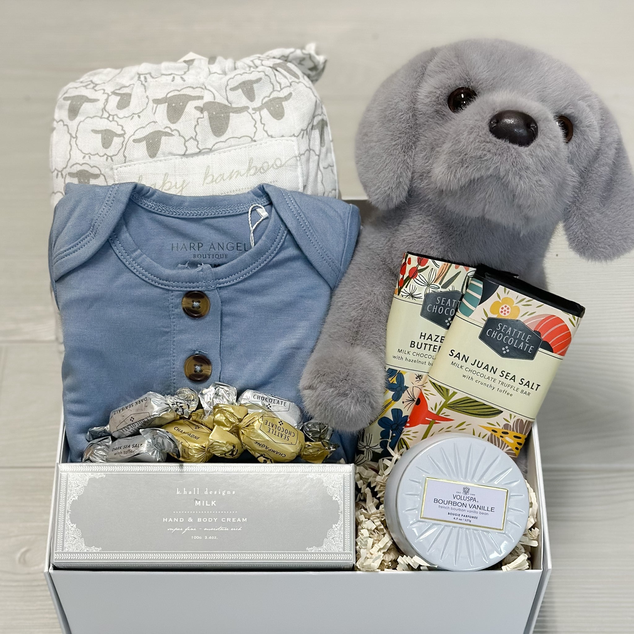 swaddle blanket, gown, stuffed dog, hand cream, candle, chocolate all collected in our snips & snails & puppy dog tails gift basket.