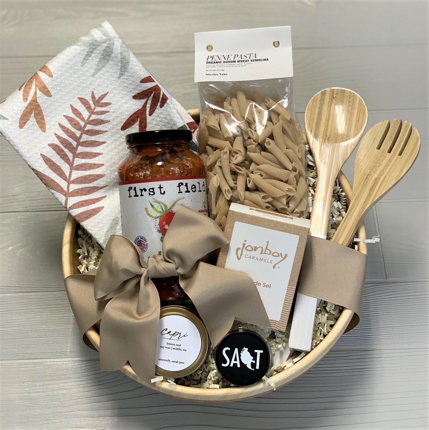 Pasta night gift basket includes pasta sauce, a kitchen towel penne pasta, serving spoons, caramel, a scented candle, and sea salt. It is packaged in a bamboo bowl.