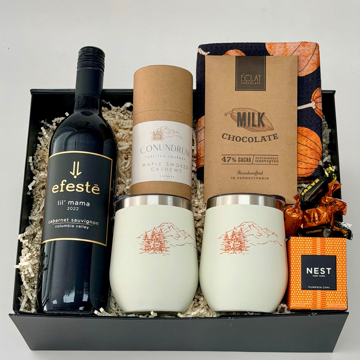 Gift box that includes a bottle of red wine, wine tumblers, cashews, chocolate, dish towel, scented candle.