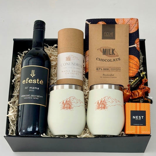 Gift box that includes a bottle of red wine, wine tumblers, cashews, chocolate, dish towel, scented candle.