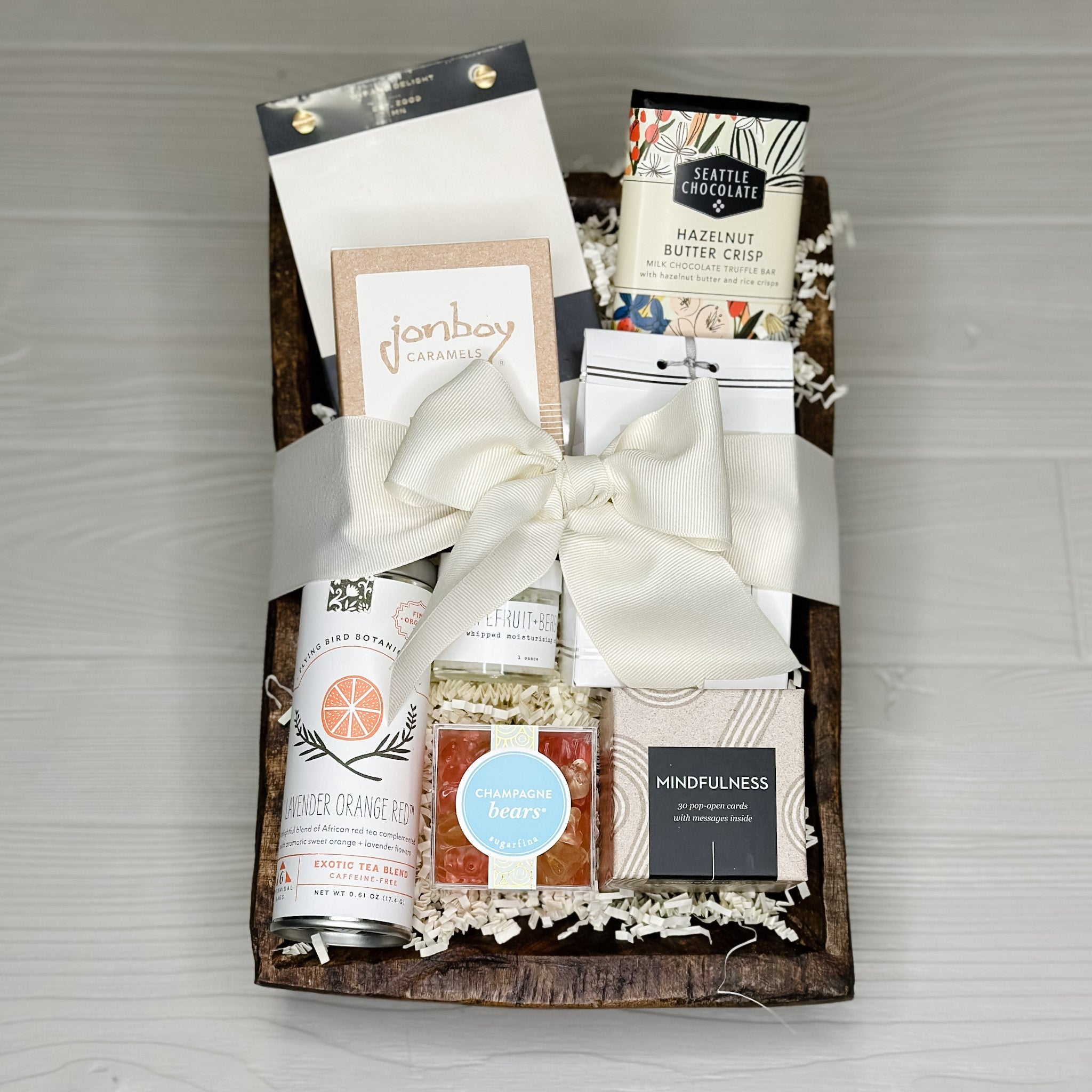 A wellness gift basket that includes organic tea, Sugarfina gummy bears, Inspirational quotes, bath salts, hand cream, caramel, chocolate, and a notepad all packaged in a wooden bowl..