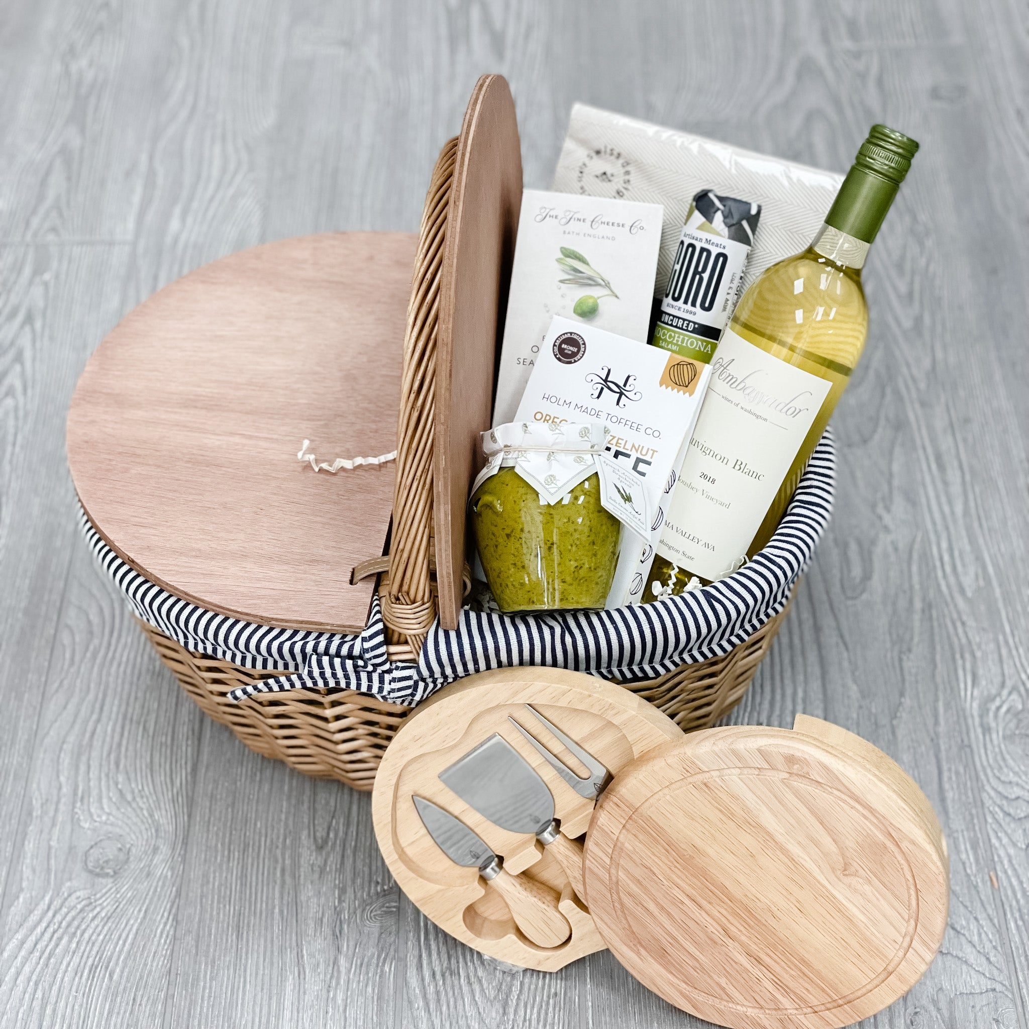 Picnic gift basket includes cheese knives, cheese board, artichoke dip, white wine, toffee, crackers, and napkins packaged in a picnic basket.